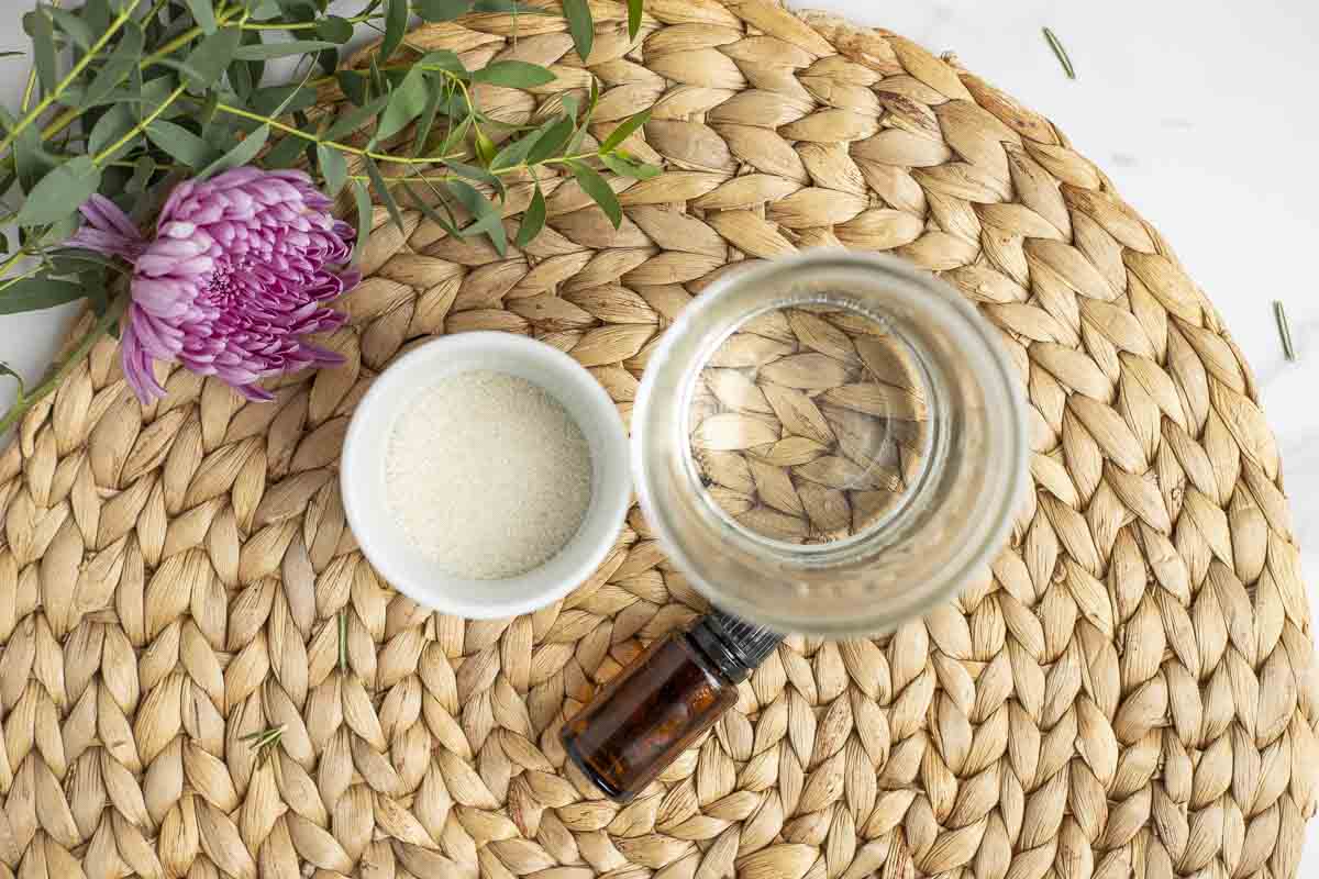small jars of sugar and water with a bottle of essential oil on a woven mat with a flower in the back corner.