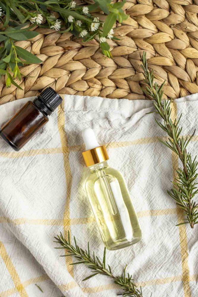 overhead picture of bottle of hair oil laying on a towel on a placemat surrounded by rosemary and an essential oil bottle