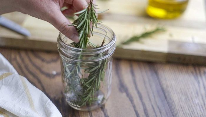 adding rosemary sprigs to a jar with a cutting board in the background
