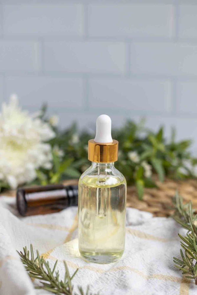 a glass bottle with dropper full of castor oil infused with rosemary on a white and yellow towel surrounded by florals and rosemary