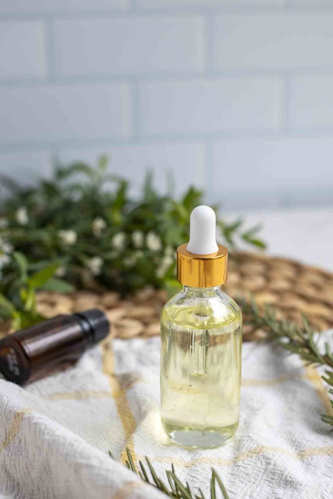 a glass dropper bottle of rosemary oil on a white and yellow towel with fresh greenery and an essential oil bottle in the background