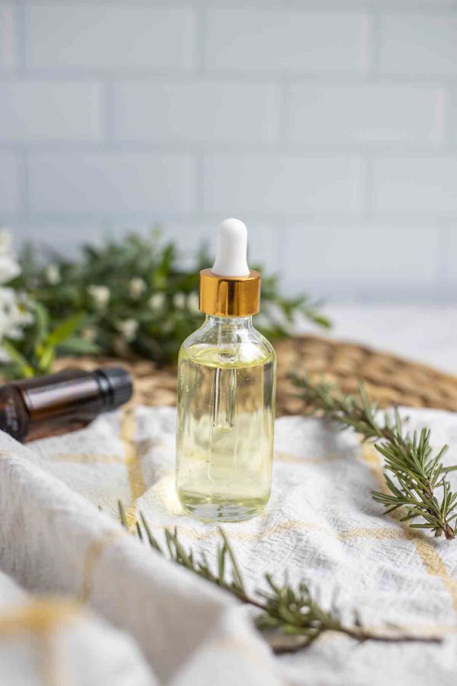 Glass bottle with dropper of rosemary hair growth oil on a white and yellow napkin with fresh sprigs of rosemary surrounding the jar. There is a small bouquet of greenery in the background