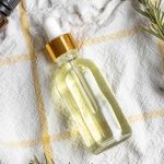overhead photo of a bottle of rosemary hair oil laying on a white and yellow plaid towel surrounded by rosemary