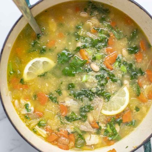 overhead photo of a dutch oven with chicken quinoa soup topped with fresh parsley and slices of lemon on a white countertop
