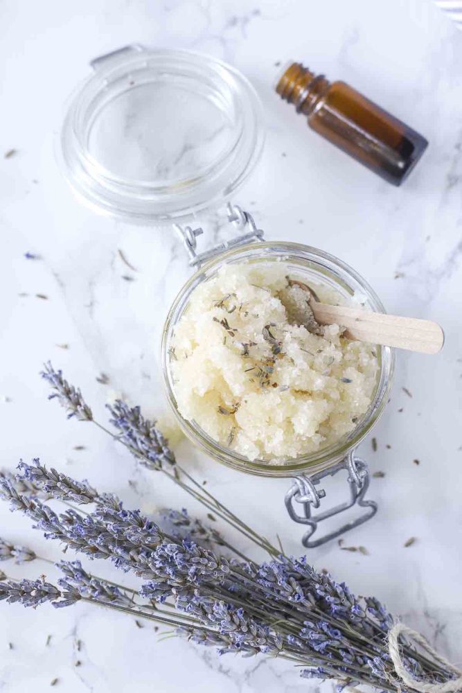 glass jar with a swing top lid filled with homemade foot scrub and topped with dried lavender. A small wooden spoon is in the jar. The jar sits on a marble counter and is surrounded by fresh lavender and an essential oil bottle