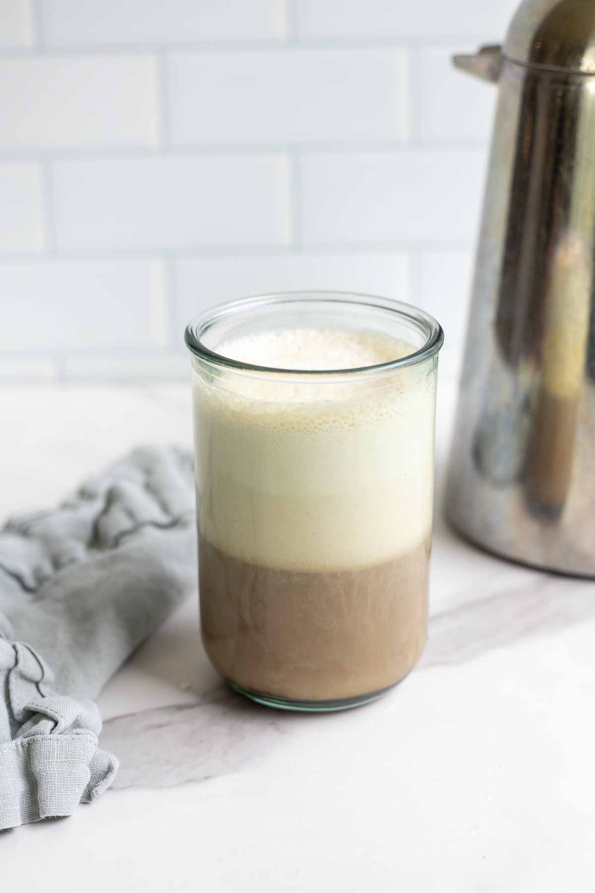 a glass of coffee topped with sweet cream cold foam on a marble countertop with a blue napkin and a metal French press in the background.