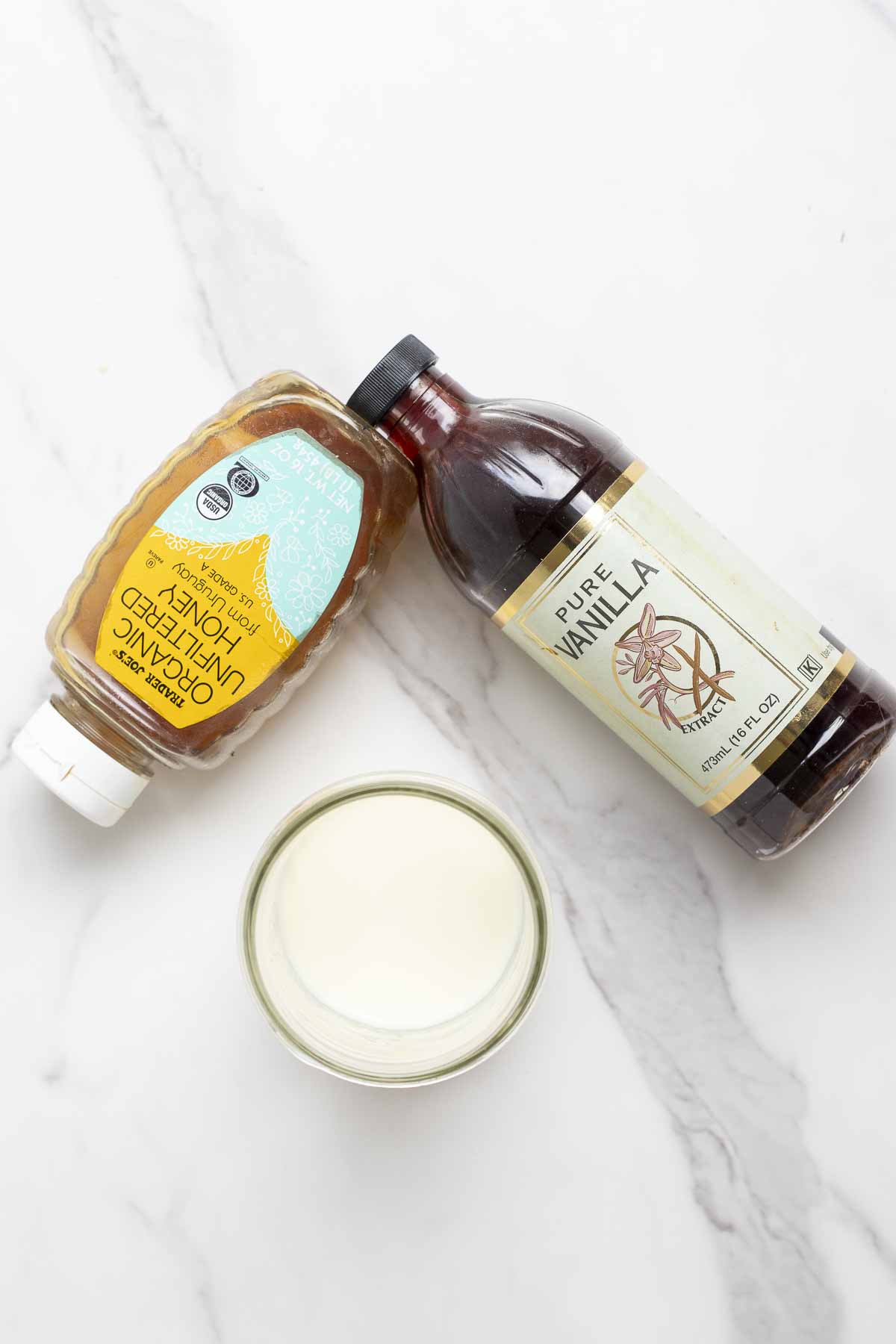 a bottle of honey, a bottle of vanilla extra, and a jar of milk on a marble counter.