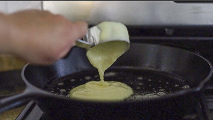 hand pouring pancake batter from a measuring scoop into a cast iron skillet