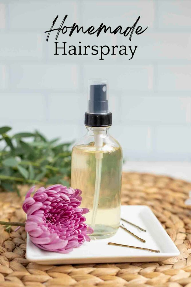 bottle of homemade hairspray on a white plate with a pink flower and bobby pins resting on the plate. The plate is on a woven place mat with greenery in the background