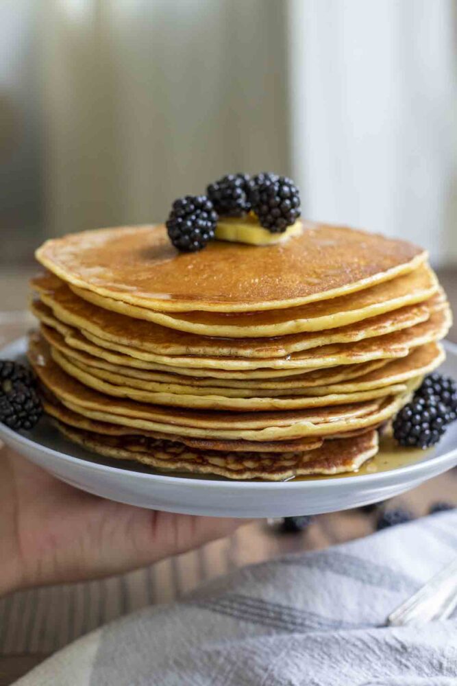 hand holding up a plate full of a stack of sourdough pancakes topped with butter and fresh blackberries