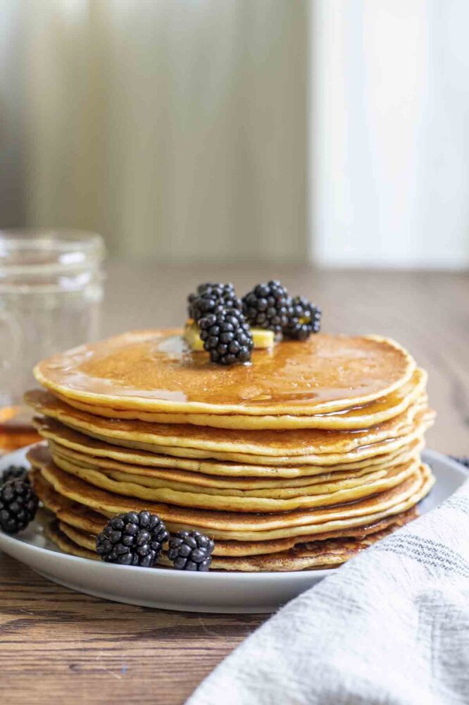 a stack of sourdough discard pancakes topped with maple syrup, blackberries, and butter on a plate with more blackberries scattered about