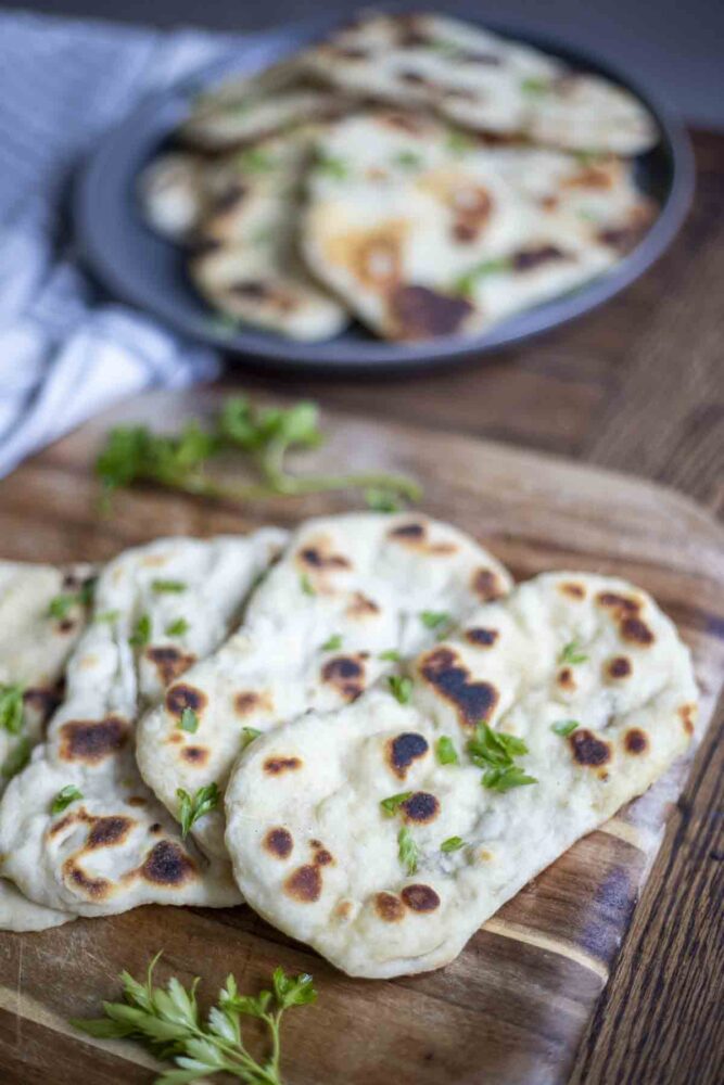 four sourdough flatbreads topped with herbs on a wood cutting board. A dark gray plate on a stripped blue and white towel has more flatbreads in the background