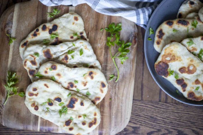 four sourdough flatbreads arranged on a wood cutting board and topped with chopped herbs. A dark gray plate in the background has more flatbreads and a stripped towel