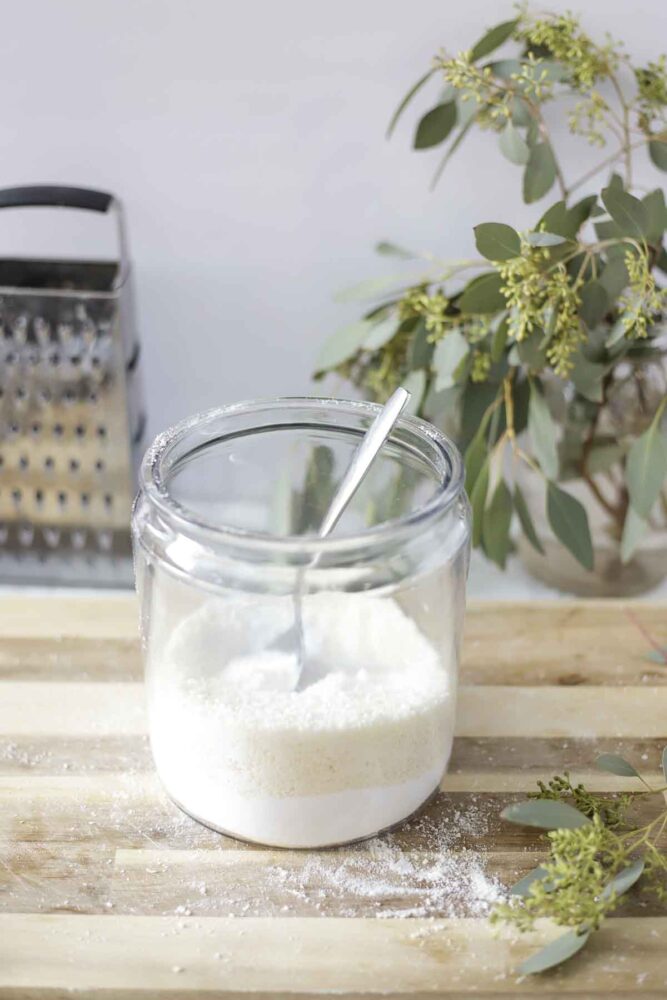 homemade laundry detergent in a jar with a spoon. The jar is on a wood cutting board with a bouquet of eucalyptus and a cheese grater in the background.