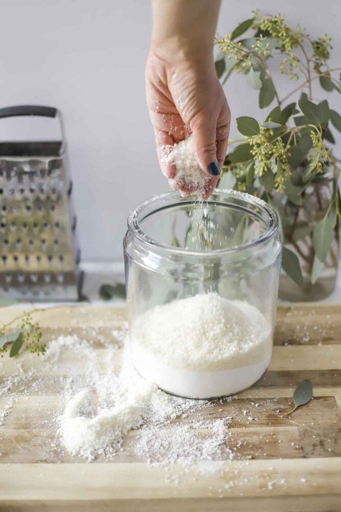 hand adding grated soap to a large class container of borax and washing soda to make homemade laundry detergent