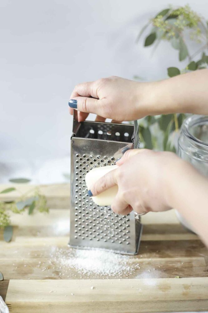 grating a bar of natural soap with a box cheese grater on a wood cutting board for homemade laundry detergent.