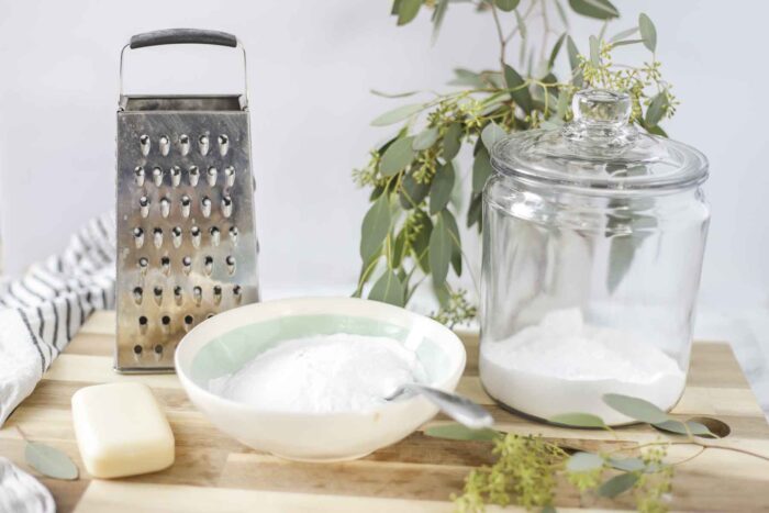 ingredients to make homemade laundry detergent on a wood cutting board with a cheese grater, and eucalyptus leaves behind