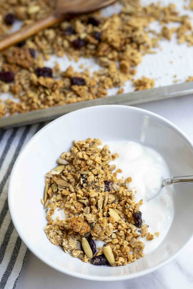 a white bowl of yogurt topped with sourdough granola. A baking sheet of more granola is in the background