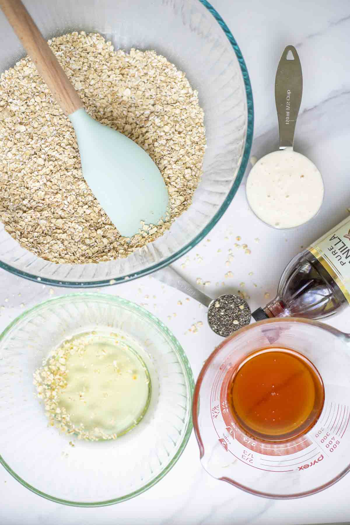 Glass bowls of oats, oil, honey, and measuring cups of sourdough starter and chia seeds and a bottle of vanilla extract are on a marble countertop.