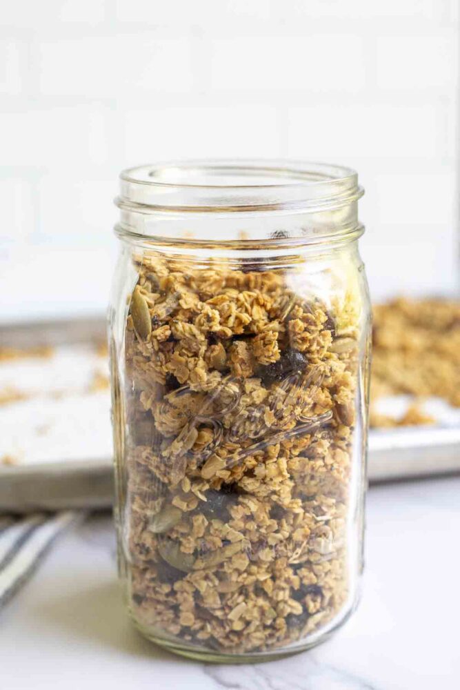 a mason jar of sourdough granola on a marble counter with a baking sheet of more granola in the background