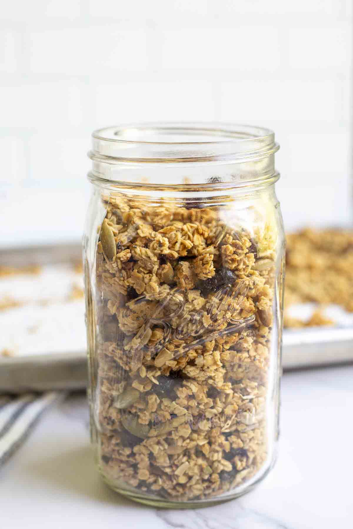 A mason jar of sourdough granola on a marble counter with a baking sheet of more granola in the background.