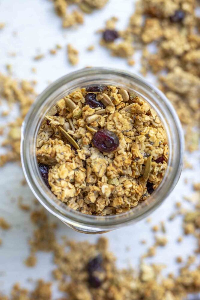 overhead photo of sourdough granola with seeds and dried cranberries in a mason jar. The jar is on a parchment lined baking sheet with more granola