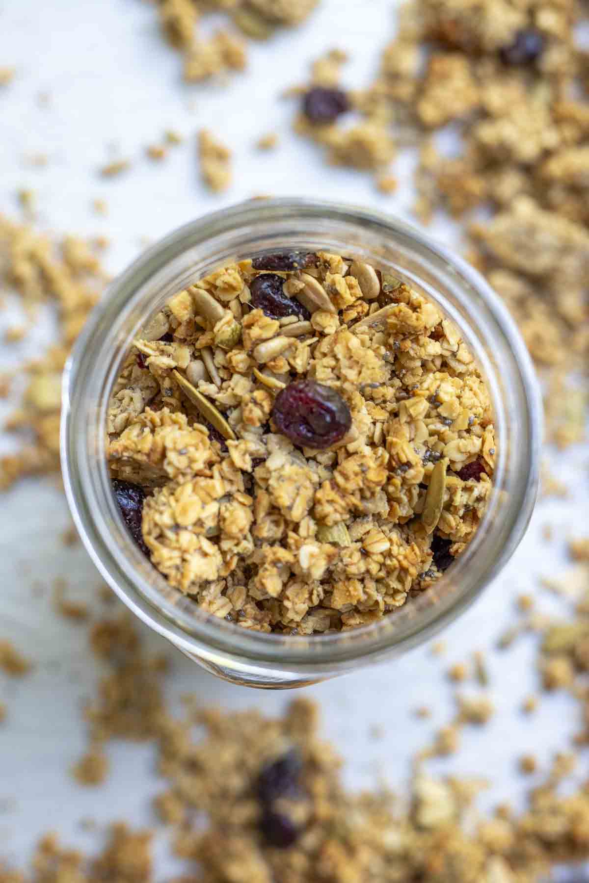 Overhead photo of sourdough granola with seeds and dried cranberries in a mason jar. The jar is on a parchment lined baking sheet with more granola.