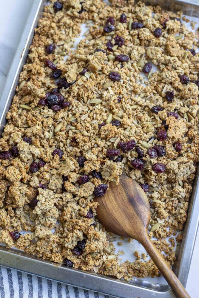 sourdough granola with sunflower seeds, pumpkin seeds, and dried cranberries on a parchment lined baking sheet with a wooden spoon