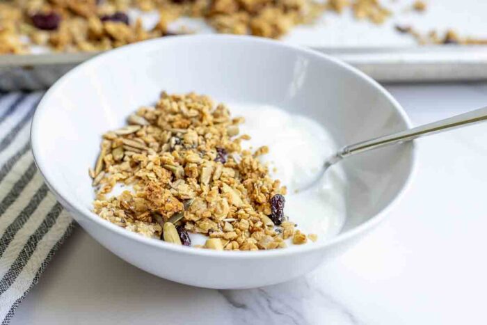 a bowl of yogurt topped with granola. A black and white stripped towel and a pan of granola are in the background