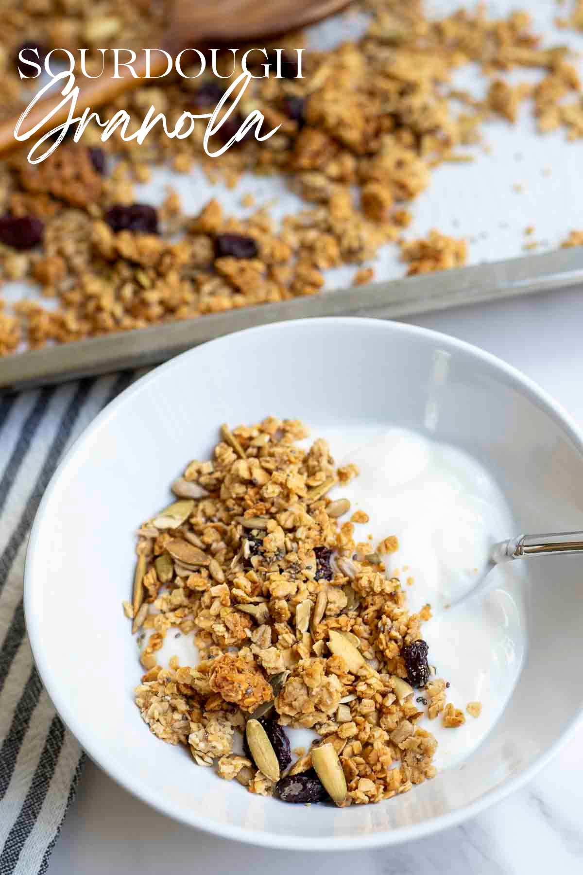 A white bowl of yogurt topped with sourdough granola. A baking sheet of more granola is in the background.