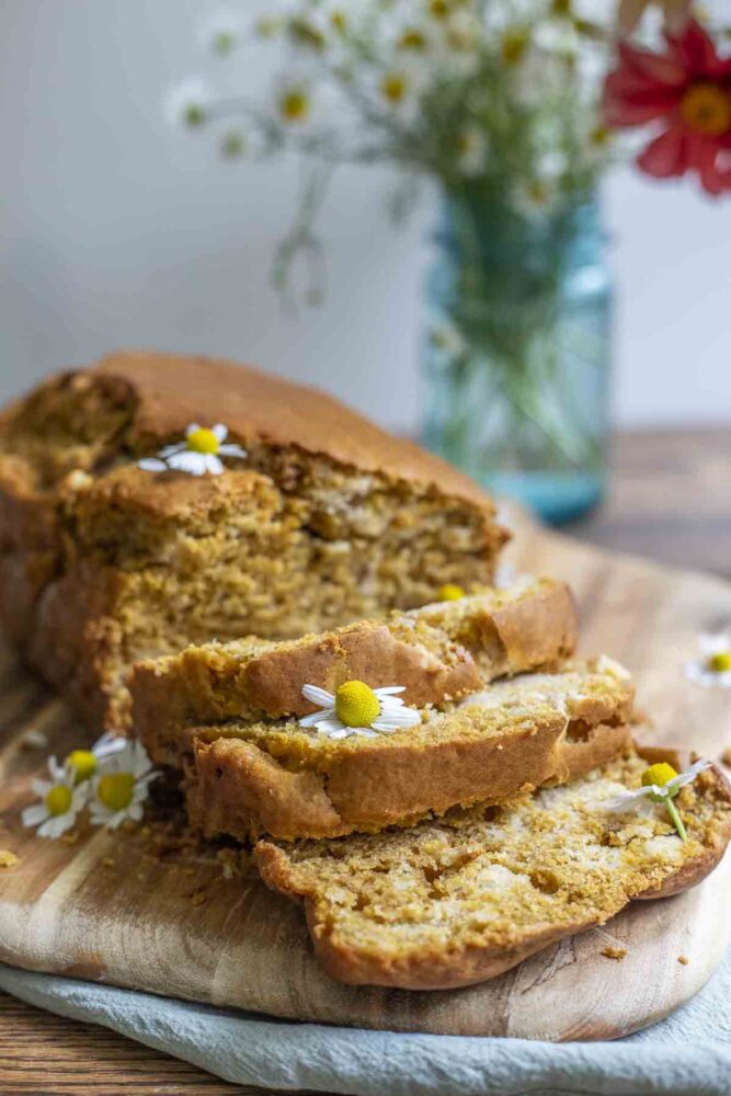 side view of a loaf of sourdough pumpkin bread sliced on a wood cutting board on top of a green towel with a vase of flowers in the background