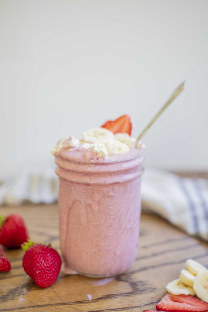 strawberry banana smoothie in a mason jar with it dripping down the sides. The smoothie is topped with fresh whipped cream, sliced strawberries and bananas. More strawberries and bananas surround the smoothie with a towel in the background