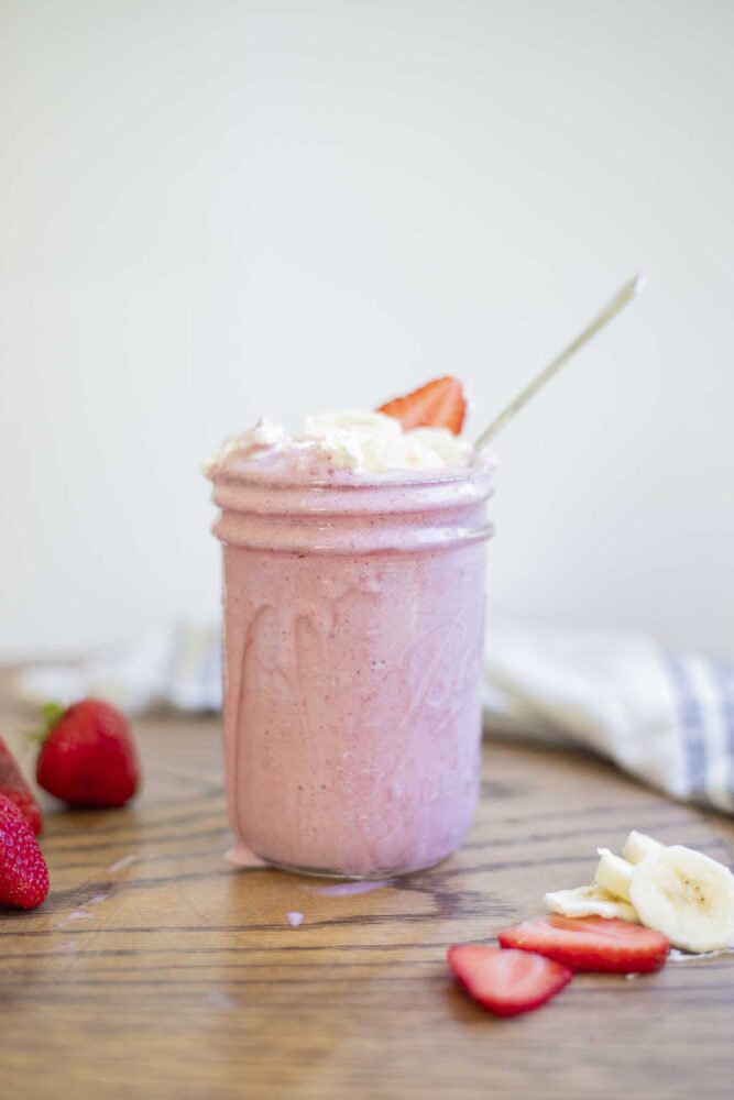 a healthy strawberry banana milkshake in a mason jar topped with whipped cream, banana slices, and strawberry slices. The jar is on a wood countertop with sliced and whole strawberries, and bananas slices surrounding.