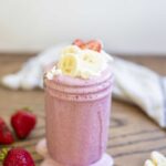 strawberry banana smoothie in a mason jar with it dripping down the sides. The smoothie is topped with fresh whipped cream, sliced strawberries and bananas. More strawberries and bananas surround the smoothie with a towel in the background