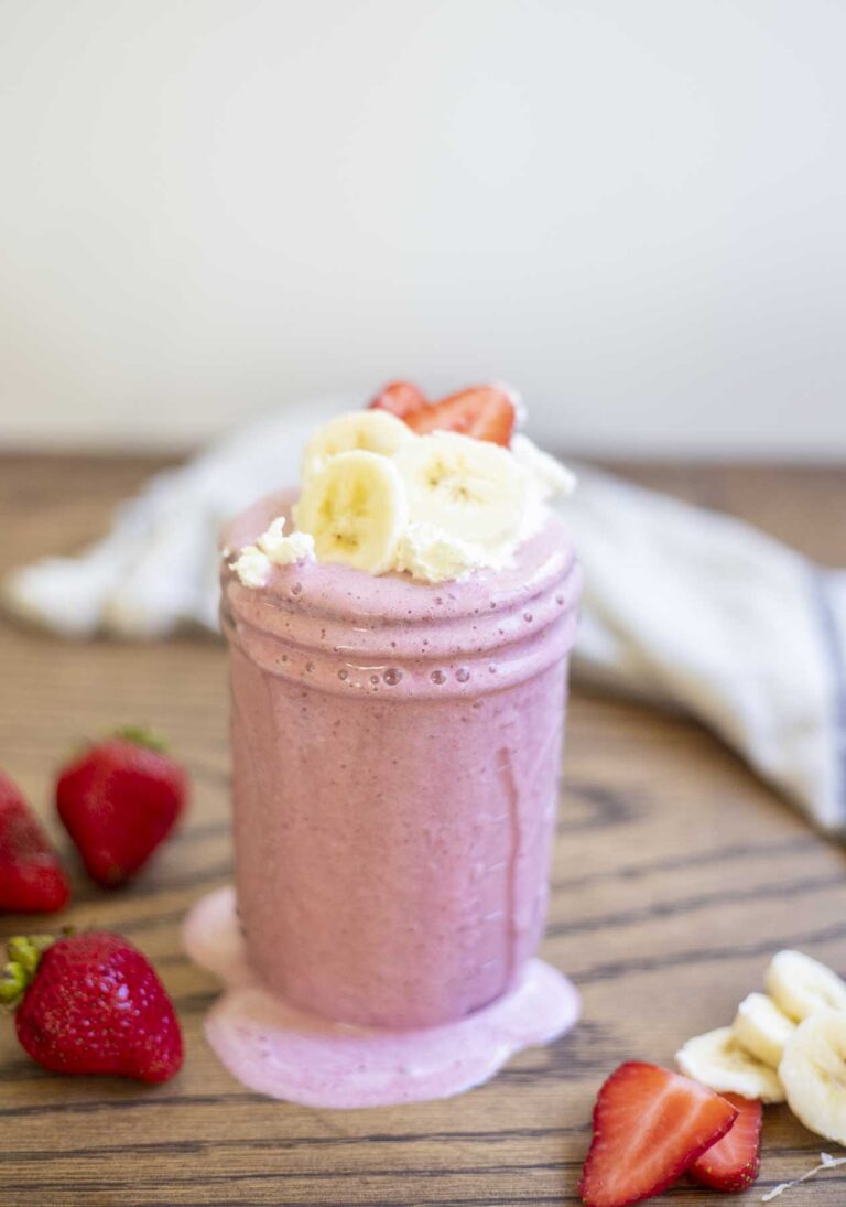 strawberry banana smoothie in a mason jar with it dripping down the sides. The smoothie is topped with fresh whipped cream, sliced strawberries and bananas. More strawberries and bananas surround the smoothie with a towel in the background