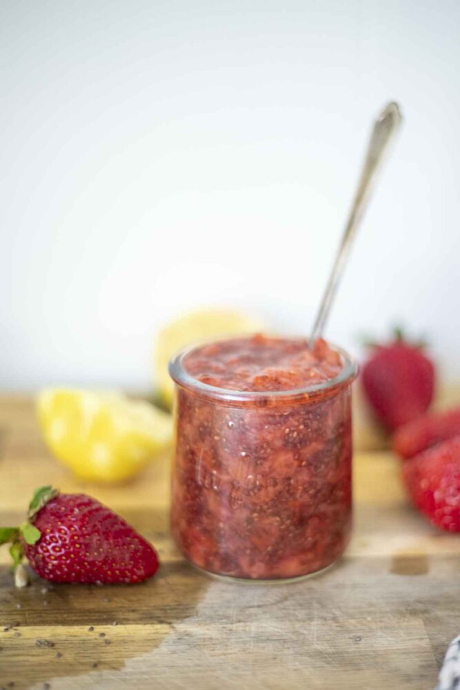 small glass jar of strawberry chia jam on a wood cutting board with a black and white towel in front and strawberries, and halved lemons in the background