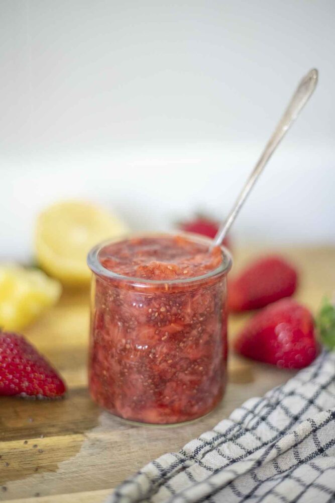 jar of strawberry chia jam on a wood cutting board surrounded by fresh strawberries and halved lemons