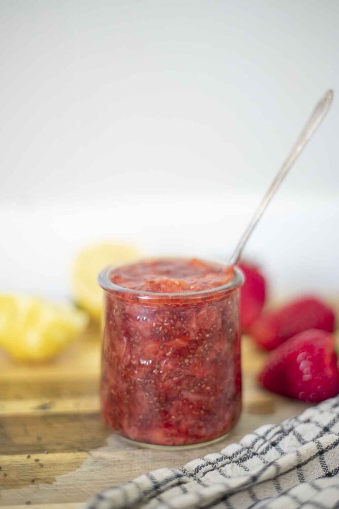 jar of strawberry chia jam on a wood cutting board surrounded by fresh strawberries and halved lemons.