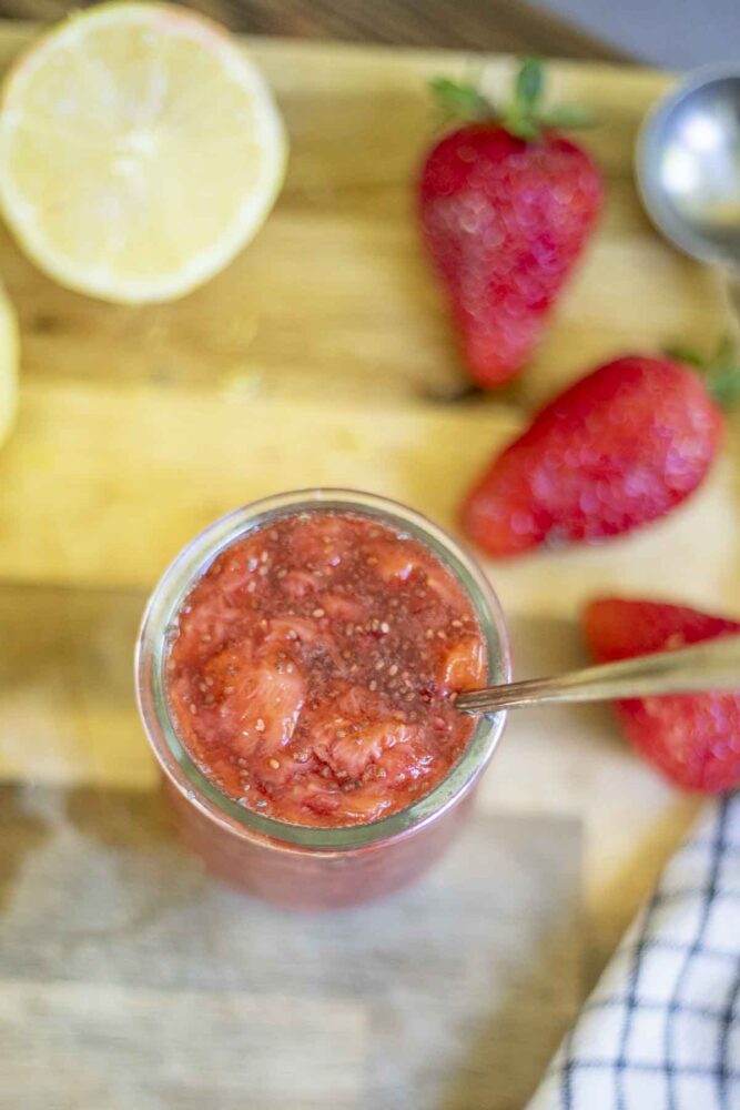 overhead photo of chia jam in a glass jar with a spoon.  A half of lemon and strawberries sit to the right
