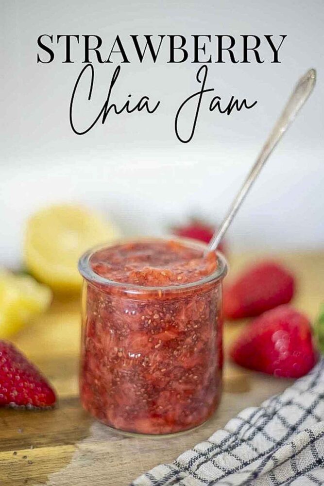 small jar filled with strawberry chia jam with a. long spoon coming out. The jar is placed on a wood countertop with a black and white towel, strawberries, and lemon.