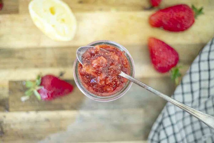 overhead photo of a spoon with chia jam on top of a jar of chia jam on wood countertop with strawberries and lemon