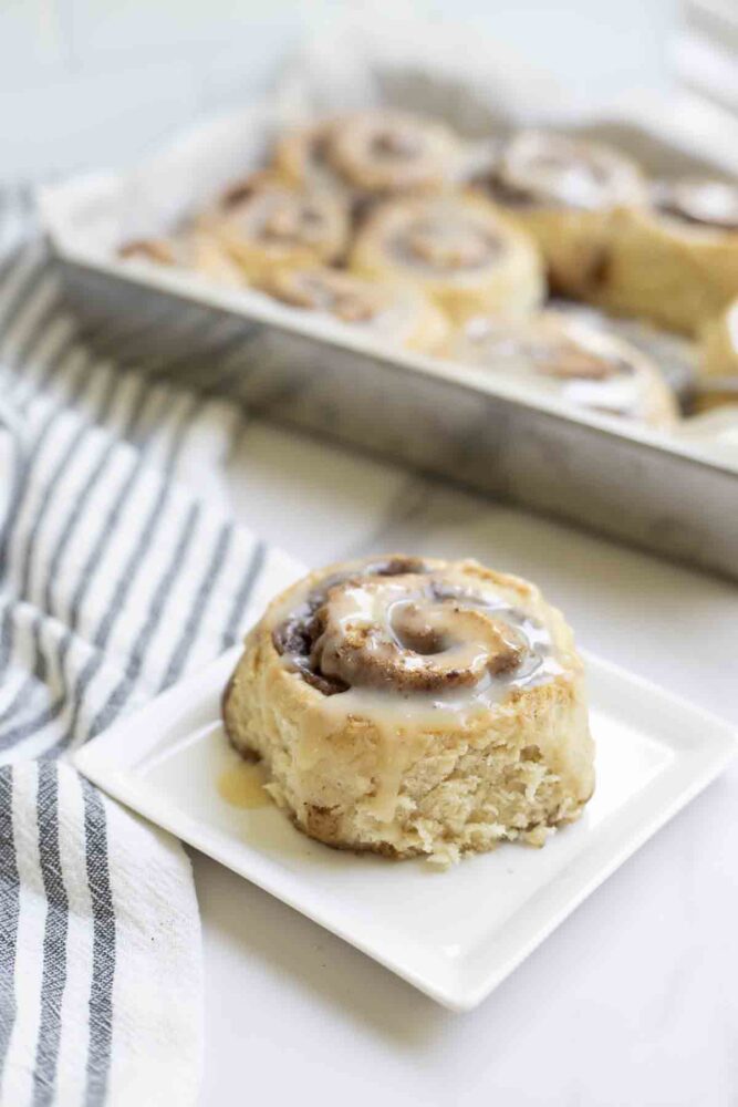 sourdough cinnamon roll on a white plate. A black and white stripped towel and a pan of cinnamon rolls is in the background