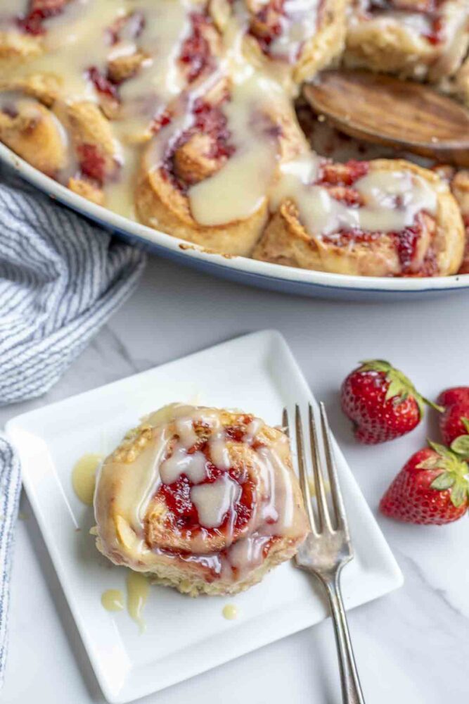 sourdough strawberry roll on a white plate with a fork on marble countertop. fresh strawberries are to the right and more rolls in a baking sheet are in the background