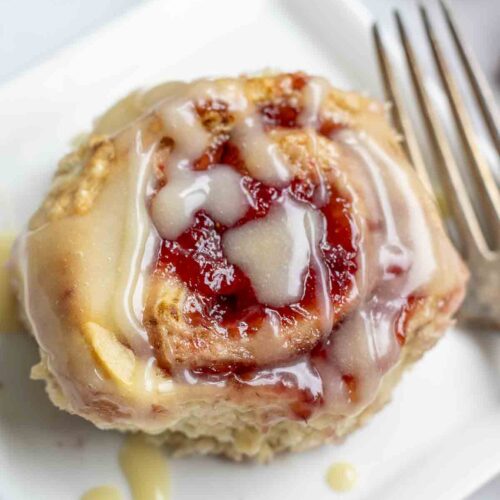 overhead photo of a sourdough strawberry sweet roll drizzled with icing on a white square plate with a antique fork to the right