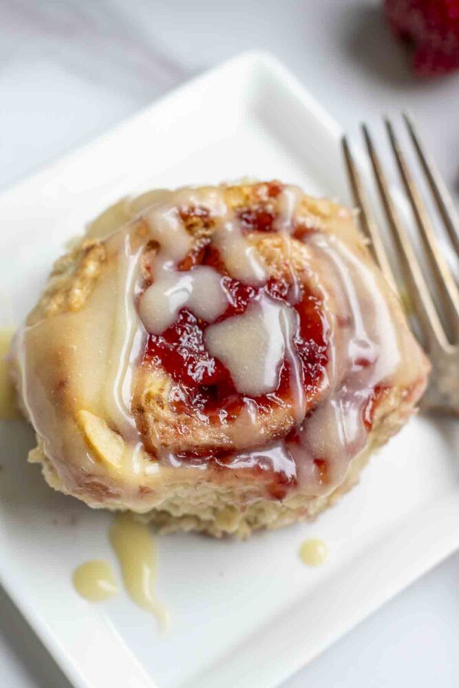 overhead photo of a sourdough strawberry sweet roll drizzled with icing on a white square plate with a antique fork to the right