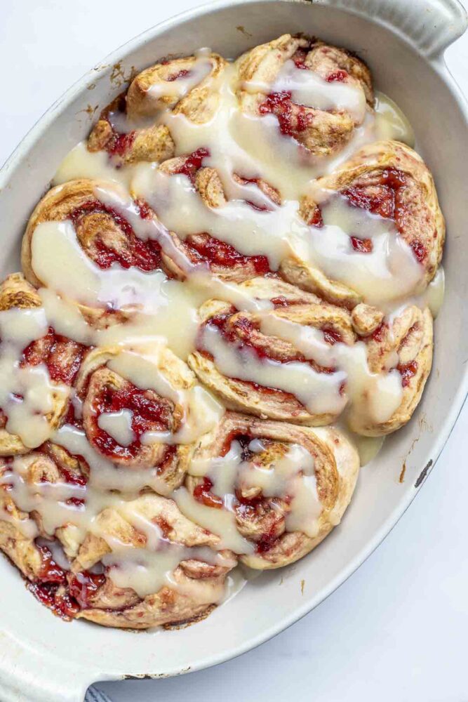 overhead photo of a oval baking dish full of sourdough strawberry rolls topped with cream cheese icing.