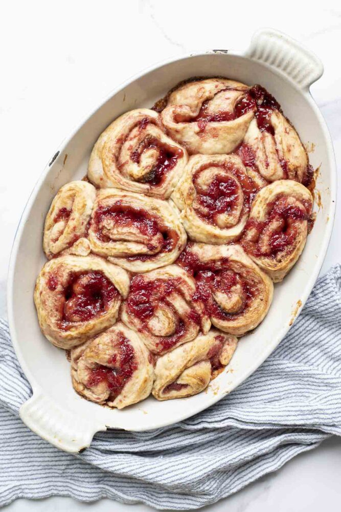 overhead photo of sourdough strawberry rolls in a oval baking dish on a marble countertop with a blue and white stripped towel