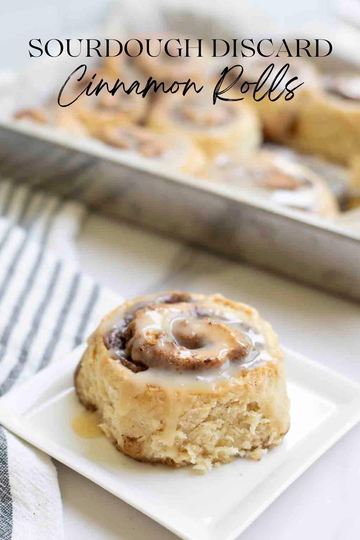 Sourdough discard cinnamon roll topped with icing on a white plate with a pan of more cinnamon rolls on a white and black stripped plate in the background