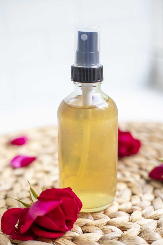 a small glass spray bottle with rose water on a woven place mat with rose petals on top