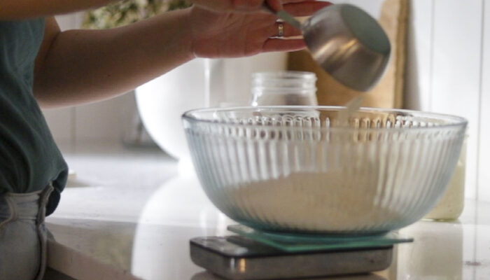 glass bowl on a kitchen scale. A person is adding flour to the bowl.